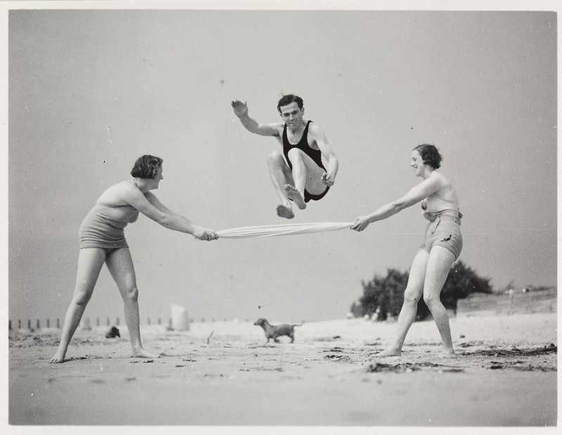A man jumping over a towel held up high by two women, on a beach.